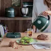 A person pouring water from a green kettle into a glass mug, with a toaster, stand mixer, avocado toast, and bread on a wooden table. Produktbild-Vorschau