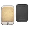 A clear plastic food storage container filled with a light brown granular substance, placed next to its dark gray lid, on a white background. Produktbild-Vorschau