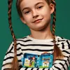 A young girl with braided hair holds up two open LEGO cubes, showing the mini-figures and accessories inside. Produktbild-Vorschau