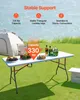 A white folding table set up outdoors on grass, with a tent and water in the background. The table is laden with food and drinks. Produktbild-Vorschau
