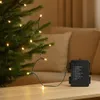A string of warm white LED lights is draped over a Christmas tree branch, with the battery pack resting on a wooden table. Produktbild-Vorschau