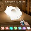 A person is holding a remote control and pointing it at a white bunny-shaped night light. Below the night light are several smaller images of the night light in different colors. Produktbild-Vorschau
