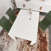 A white dining table with green chairs around it, on a patterned rug. Produktbild-Vorschau