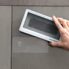 A hand holding a white waterproof phone case with a dark object inside, attached to a tiled wall. Podgląd zdjęcia produktu