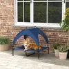 A dog is resting on an elevated dog bed with a canopy, placed on a patio next to a brick wall and potted plants. Anteprima dell’immagine prodotto