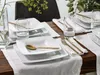 A table setting with white square plates, gold cutlery, and glasses of water, arranged on a wooden table with a white runner. Náhľad obrázku produktu
