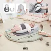 A baby is lying on a round mat with a baby lounger. There are also some toys and a book on the mat. In the background, there is a washing machine with a baby lounger inside. Produktbild-Vorschau