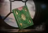 A green and gold deck of cards is displayed in front of a circular window with a cloudy sky visible. Náhled obrázku produktu