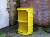 A yellow barrel shelf unit with two shelves is placed outdoors next to a brick wall and a white door. Podgląd zdjęcia produktu