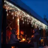 Three people sitting around a table outside a wooden cabin at night, illuminated by icicle lights and a candle. Produktbild-Vorschau