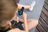 A person is using a massage gun on their leg while sitting on a yoga mat. Produktbild-Vorschau