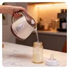 A hand is pouring water from a beige and brown electric kettle into a baby bottle, with a bottle cap next to it. Podgląd zdjęcia produktu