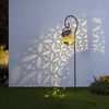A solar-powered watering can garden light casting decorative shadows on a wall and illuminating the ground with fairy lights. Produktbild-Vorschau