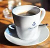 A white coffee cup with the Dallmayr logo sits on a saucer on a wooden table. Podgląd zdjęcia produktu