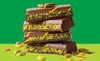 A stack of chocolate bars filled with pistachio paste, with loose pistachios scattered around the bottom. Produktbild-Vorschau