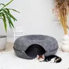 A kitten is resting in a round, zippered cat bed placed on a rug. A plant and a decorative vase with dried flowers are in the background. Produktbild-Vorschau