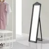 A full-length mirror with a dark gray frame stands on a light gray floor in a room with light gray walls. To the left, a white shelf unit holds shoes and a book, and a clothing rack displays a pink dress and a white shirt.