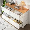 A white and wood sideboard with open drawers filled with various items, placed in a room with a rug and plants. Anteprima dell’immagine prodotto