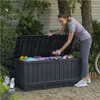 A woman is putting a pink dumbbell into a large dark gray storage box in a garden setting. Podgląd zdjęcia produktu