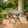 A square folding table with a wicker-like top and black metal legs is placed on a wooden deck. The table is surrounded by potted plants and a garden with trees and flowers in the background. Produktbild-Vorschau