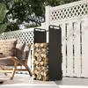 A black metal firewood rack filled with logs is placed on a patio next to a chair and a white lattice fence. Produktbild-Vorschau