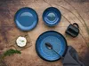 A collection of blue ceramic dishes and a black mug arranged on a wooden surface, with a small piece of bread topped with cream cheese and dill on a wooden cutting board. Náhľad obrázku produktu