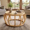 A round wooden coffee table with a curved frame base, placed on a textured rug in a living room setting. A vase of flowers, a stack of books, and a cup of coffee are on the table. Two armchairs are visible on either side, and a large window with a view of  Anteprima dell’immagine prodotto