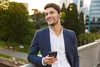 A man in a suit is holding a smartphone and wearing earbuds, standing on a bridge with a city park in the background. Podgląd zdjęcia produktu
