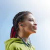 A young woman wearing headphones and a green hoodie, looking up and to the right with a slight smile. Anteprima dell’immagine prodotto