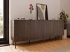 A modern sideboard with a fluted front is placed in a well-lit room with wooden floors. A framed artwork and a vase with flowers are on top of the sideboard. Produktbild-Vorschau