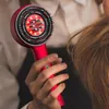 Rote Haartrocknerbürste mit schwarzen Borsten und glühendem roten Zentrum, gehalten von einer Hand in der Nähe von hellbraunem Haar Produktbild-Vorschau