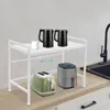 A white kitchen shelf unit holding two black kettles and a white mug on the top shelf, and a green toaster and a gray air fryer on the countertop below. A kitchen sink and faucet are visible to the right. Náhled obrázku produktu