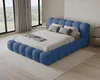 A blue upholstered bed with pillows and a blanket is shown in a bedroom setting with large windows and curtains. Voir les images du produit