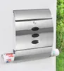 A stainless steel mailbox mounted on a white wall, with greenery in the background. A rolled newspaper is visible in the lower compartment. Produktbild-Vorschau