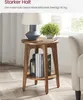 A round wooden side table with a lamp, a plant, and books on it, placed in a room with a chair and a window in the background. Produktbild-Vorschau