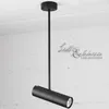 A black cylindrical pendant light hanging from a black rod against a textured white wall. Podgląd zdjęcia produktu