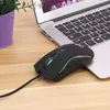 A black wired gaming mouse with RGB lighting is placed on a wooden desk next to a laptop. Produktbild-Vorschau