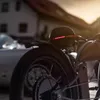 Close-up of the rear of a black bicycle helmet with red LED lights, mounted on a bicycle, with a classic car in the background. Náhled obrázku produktu