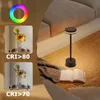 A modern black LED table lamp is placed on a textured rug next to a sofa, with an open book on the rug. The image also includes inset panels showing sunflowers in a vase under different CRI lighting conditions and a color spectrum graphic. Náhľad obrázku produktu