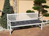 A white wooden bench with a dark gray cushion is placed outdoors on a brick patio, next to a tall evergreen tree and a decorative tree. Náhled obrázku produktu