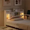 A close-up view of a white bed with a headboard featuring shelves and integrated lighting, showcasing decorative items and books. Náhled obrázku produktu