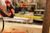 A hand is holding a tape measure to measure a piece of wood in a workshop. Produktbild-Vorschau