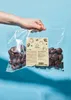 A hand holding a clear plastic bag filled with dark brown plum-shaped candies, with a product label in the center. Produktbild-Vorschau