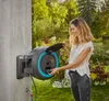 A woman is using a wall-mounted hose reel in a garden. Voir les images du produit