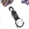 A close-up shot of the back of a black carabiner watch with a silver backplate, set against a white background with blurred pink and purple flowers in the upper left and upper right corners. Produktbild-Vorschau