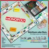 A top-down view of a Monopoly board game, laid out on a table. The game is partially set up with money, cards, and tokens visible. Produktbild-Vorschau