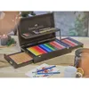 A wooden art set case with colored pencils and drawing supplies is open on a table, with a candle and a mug nearby. Anteprima dell’immagine prodotto