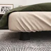 Close-up of a bed with a green throw blanket and a patterned bed skirt, resting on black furniture risers on a striped rug. Produktbild-Vorschau
