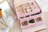 An open pink jewelry box filled with various jewelry items, including watches, rings, earrings, and necklaces. Makeup brushes and a perfume bottle are visible to the left. Náhled obrázku produktu