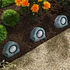 Solar-powered rock lights are placed in a garden bed with flowers and gravel. Produktbild-Vorschau
